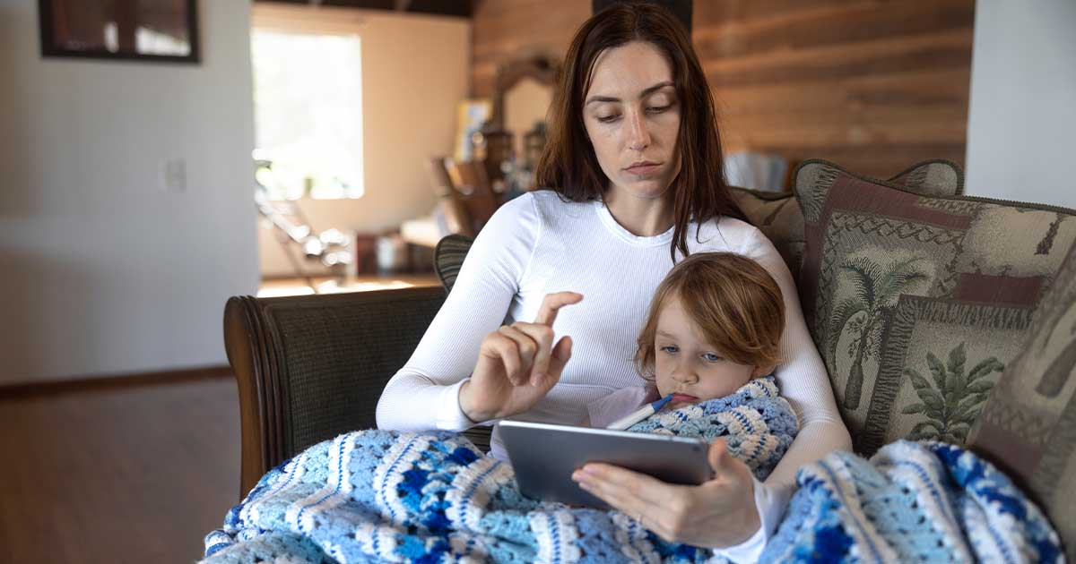 Eine Mutter sitzt mit ihrem kranken Kind auf dem Schoß auf der Couch, während die Mutter ein Tablet in der Hand hält. Das Kind hat ein Fieberthermometer im Mund.