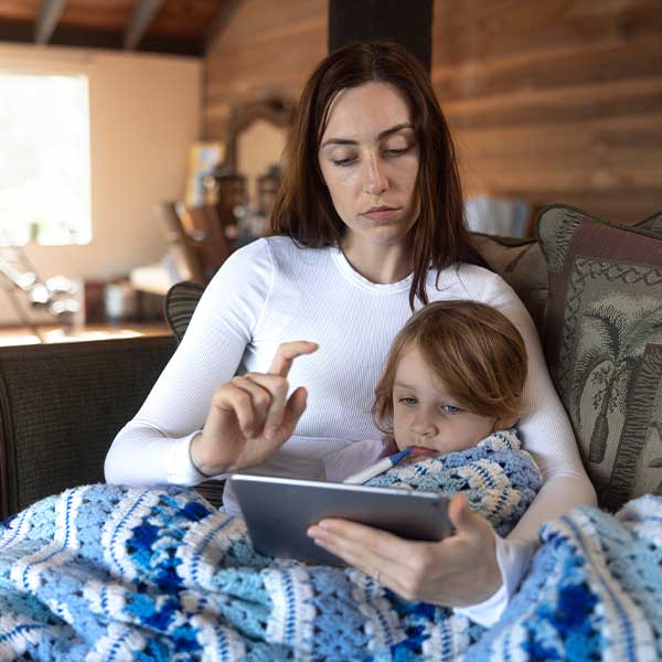 Eine Mutter sitzt mit ihrem kranken Kind auf dem Schoß auf der Couch, während die Mutter ein Tablet in der Hand hält. Das Kind hat ein Fieberthermometer im Mund.