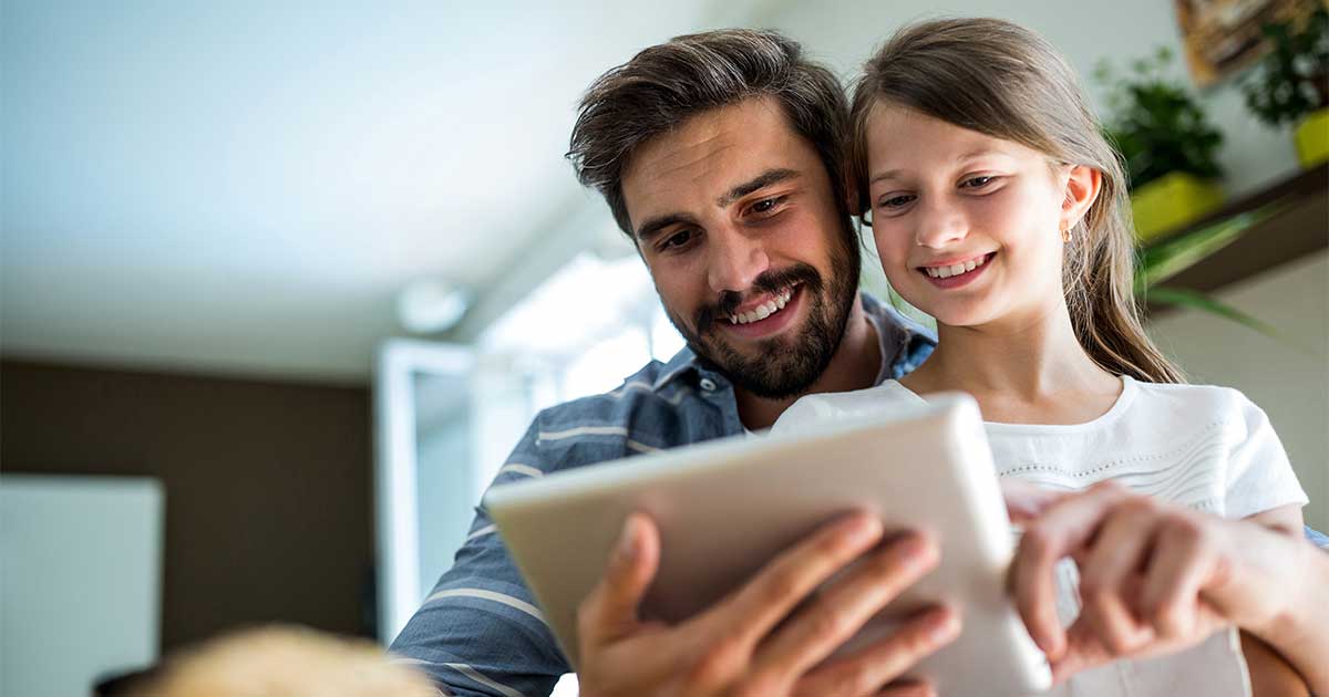 Ein Vater schaut gemeinsam mit seiner Tochter auf ein Tablet und schreiben der Lehrkraft der Tochter.