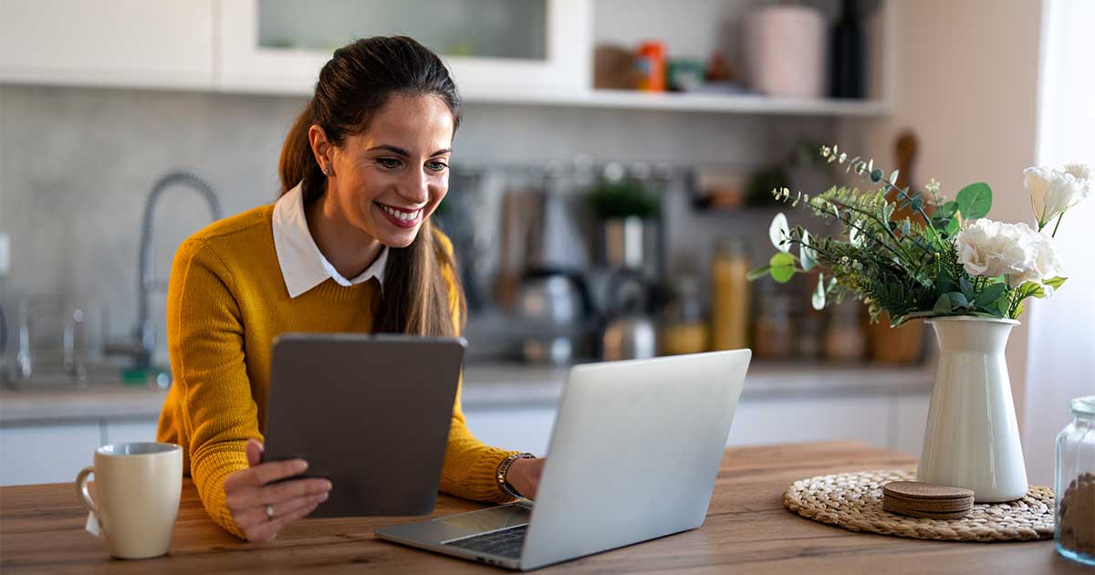 Eine Frau im gelben Pullover sitzt in ihrer Küche an einem Holztisch. Vor ihr steht ein Laptop auf den sie schaut. In ihrer rechten Hand hält sie ein Tablet neben den Laptop.