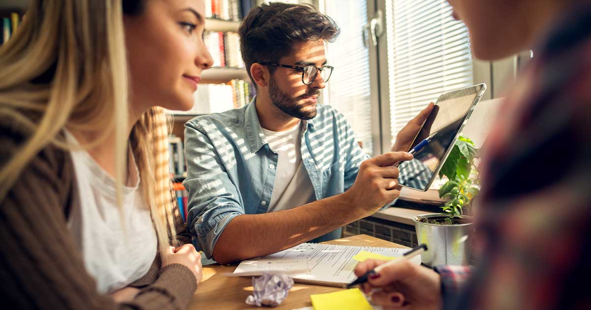 Eine Gruppe von jungen Lehrkräften sitzen an einem Tisch mit Blöcken und Stiften und machen sich Notizen. Der junge Lehrer zeigt etwas auf dem Tablet.