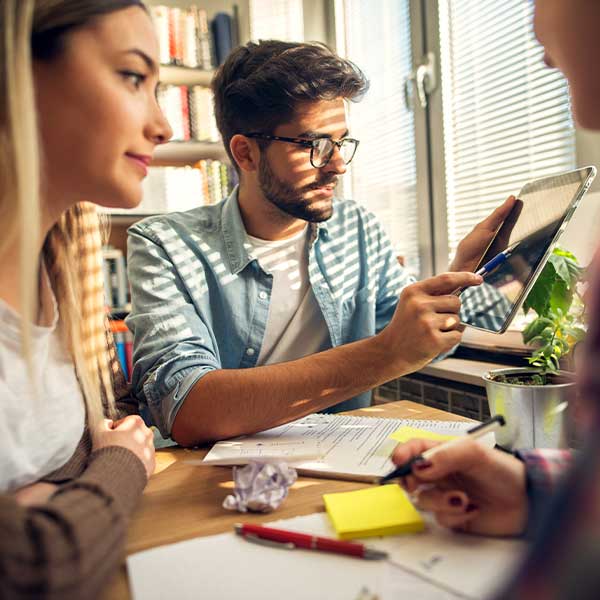 Eine Gruppe von jungen Lehrkräften sitzen an einem Tisch mit Blöcken und Stiften und machen sich Notizen. Der junge Lehrer zeigt etwas auf dem Tablet.