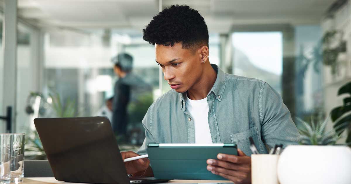 Ein junger Mann am Laptop hält ein Tablet in der Hand