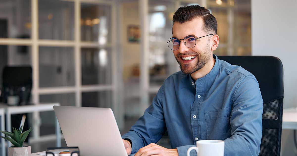 Ein junger Mann mit Brille sitzt in einem modernen Büro an einem Schreibtisch vor seinem Laptop und lächelt.