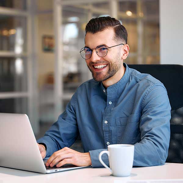 Ein junger Mann mit Brille sitzt in einem modernen Büro an einem Schreibtisch vor seinem Laptop und lächelt.