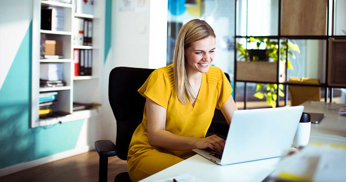 Eine Frau im gelben Kleid sitzt in einem modernen Büro am Schreibtisch an ihrem Laptop.