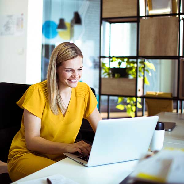 Eine Frau im gelben Kleid sitzt in einem modernen Büro am Schreibtisch an ihrem Laptop.